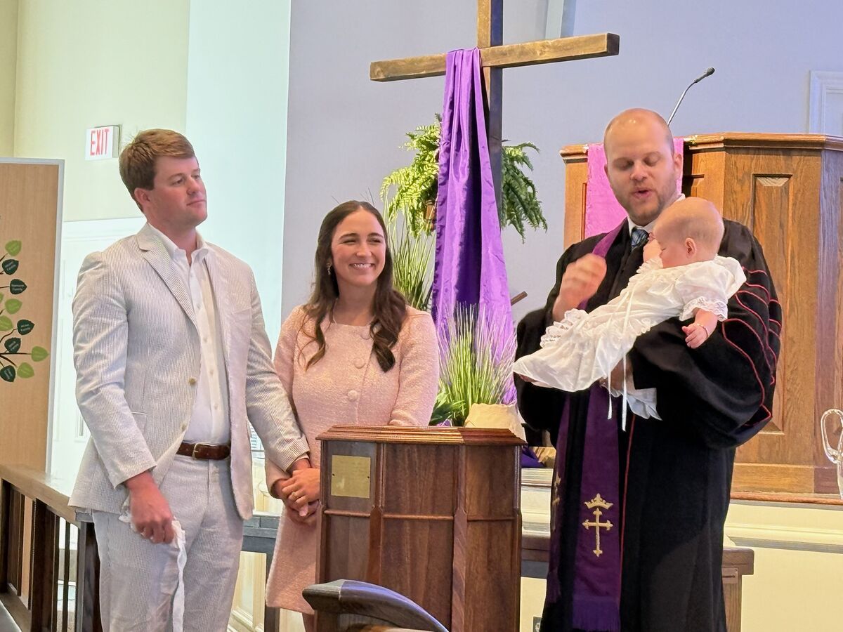 A pastor holding an infant in a christening gown at the font while her parents look on
